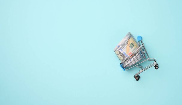 carrello della spesa in miniatura con una pila di dollari americani di carta su sfondo azzurro. Risparmio, vendita, spazio di copia - Foto, immagini