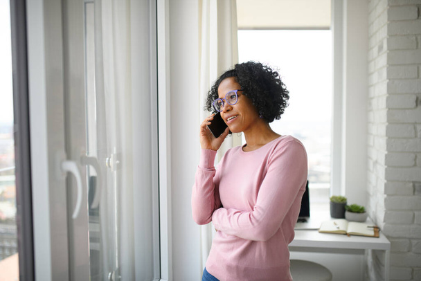 Portrait of happy mature woman making a phone call indoors, looking out of window. - Fotoğraf, Görsel