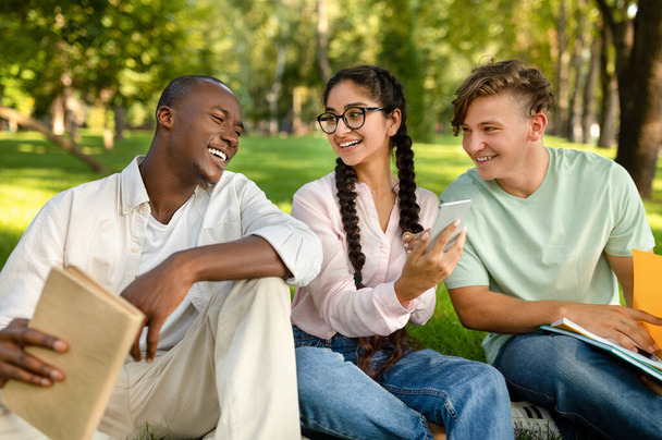 Diversos estudiantes amigos usando smartphone con nueva aplicación educativa, sentados en el campus universitario juntos - Foto, Imagen