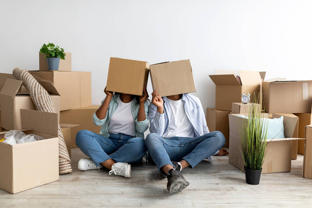 Épouses afro-américaines ludiques avec des boîtes en carton sur les têtes, assises sur le sol et célébrant le jour du déménagement, espace de copie - Photo, image