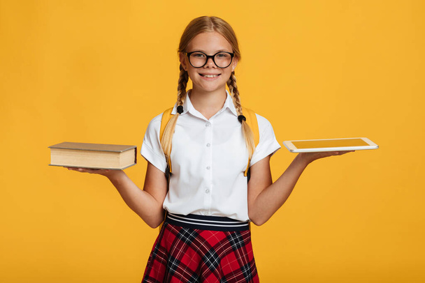 Χαρούμενη νεαρή καυκάσια μαθήτρια με κοτσιδάκια, σακίδιο πλάτης σε γυαλιά κρατήσει το βιβλίο και το tablet και επιλέγει - Φωτογραφία, εικόνα