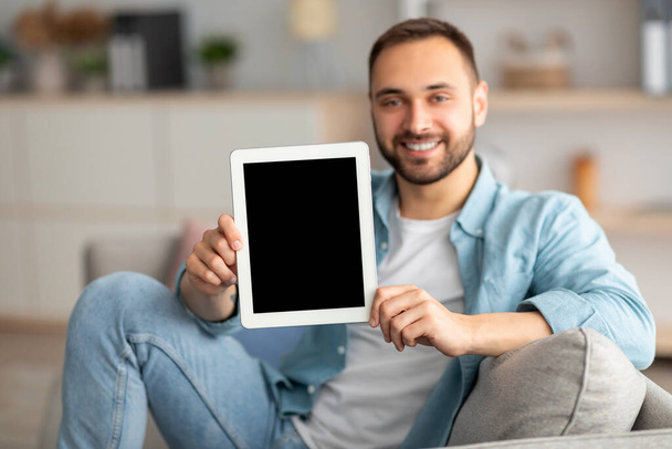 Glücklicher junger Mann zeigt Tablet-Computer mit Attrappe für Website oder Anwendung auf dem Bildschirm, drinnen - Foto, Bild