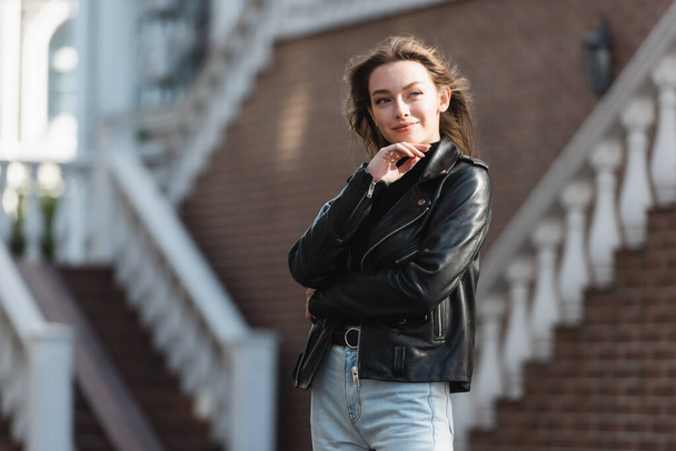 gelukkig jong vrouw in zwart lederen jas staan op stedelijke straat  - Foto, afbeelding