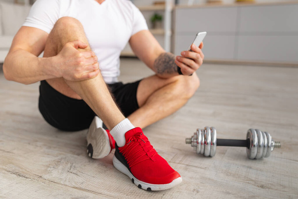 Seguimiento de la actividad del cuerpo. Hombre maduro con smartphone, sentado en el suelo con mancuernas cerca, haciendo ejercicio en la sala de estar - Foto, Imagen