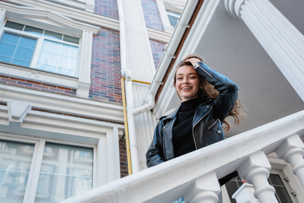 niski kąt widzenia pozytywnej młodej kobiety w skórzanej kurtce uśmiechniętej na miejskiej ulicy w Europie  - Zdjęcie, obraz