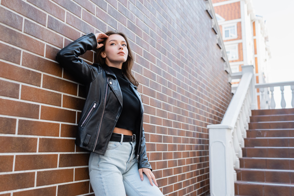 jovem sonhadora em jaqueta de couro e jeans de pé perto da parede de tijolo  - Foto, Imagem
