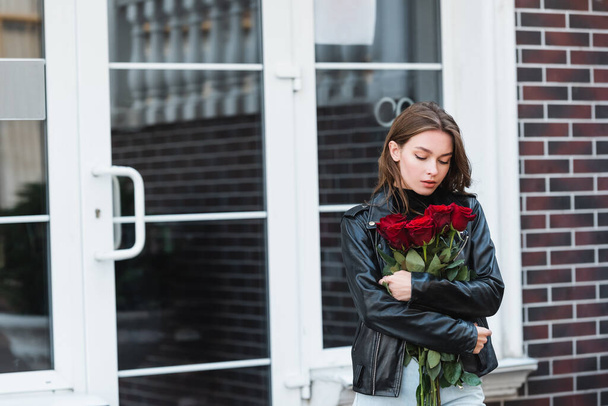 young woman in leather jacket looking at red roses on urban street in europe  - Foto, Bild