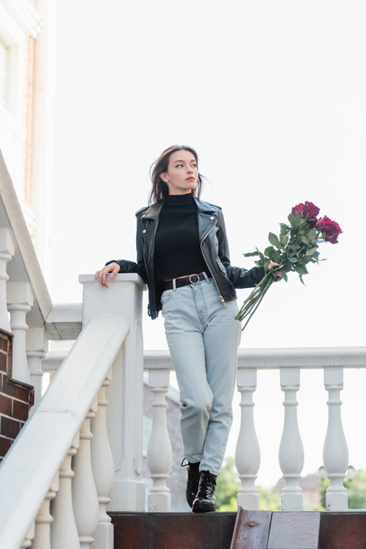 longitud completa de la mujer bonita en chaqueta de cuero negro y jeans que sostienen ramo de rosas en la calle urbana en Europa  - Foto, Imagen
