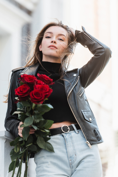 pretty woman in black leather jacket and jeans holding bouquet of roses on urban street - Foto, Imagen