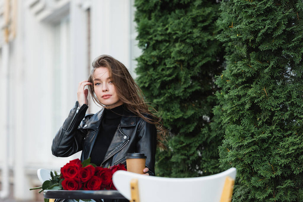 pretty woman in leather jacket holding paper cup near bouquet of red roses on coffee table  - 写真・画像