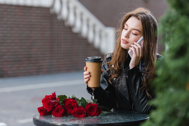 pretty woman in leather jacket holding paper cup and talking on smartphone near bouquet of red roses - Foto, immagini