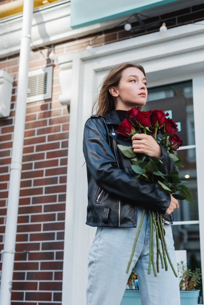 young stylish woman in leather jacket holding red roses on urban street in europe  - Fotoğraf, Görsel