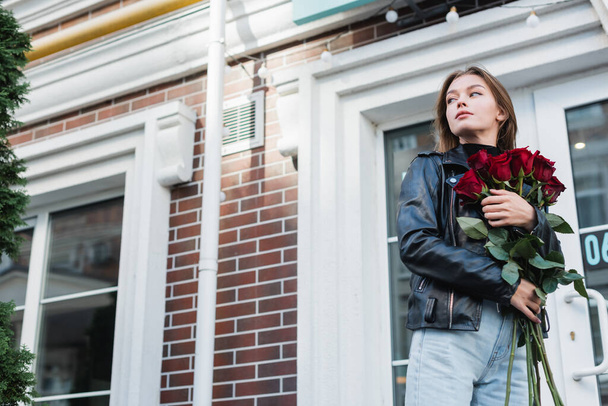 young and trendy woman in leather jacket holding red roses on urban street in europe  - Foto, Imagen