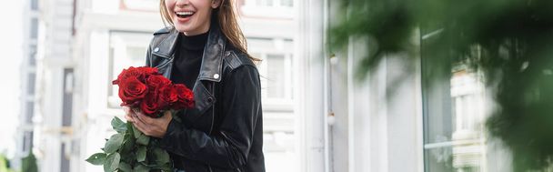 cropped view of young happy woman in leather jacket holding red roses on urban street in europe, banner - Foto, Imagem