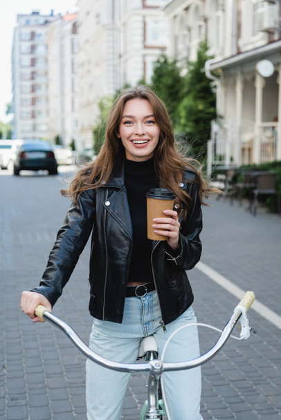 happy woman holding paper cup while riding bicycle on urban street of europe - Foto, Imagem