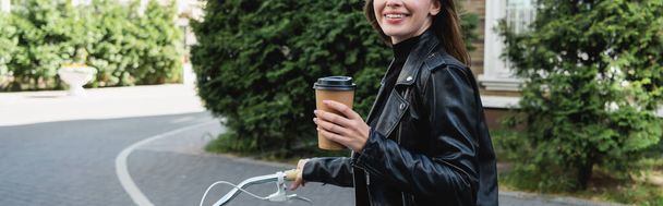 vista ritagliata della donna felice che tiene in mano la tazza di carta mentre va in bicicletta sulla strada urbana d'Europa, banner - Foto, immagini