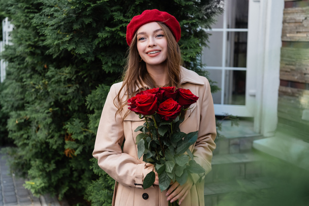 joyful woman in trench coat and red beret holding roses near building  - Valokuva, kuva