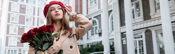 young woman in trench coat holding red roses and adjusting beret on urban street of europe, banner - Foto, Imagem