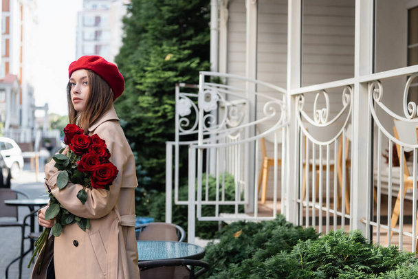 Trençkotlu ve bereli genç ve güzel bir kadın Avrupa kentsel caddesinde kırmızı güller tutuyor.  - Fotoğraf, Görsel