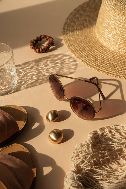 Luxury bohemian aesthetic fashion composition. Crystal glass with sunlight shadows, sunglasses, slippers, women's straw hat, string bag, gold earrings on neutral beige background - Foto, Imagem