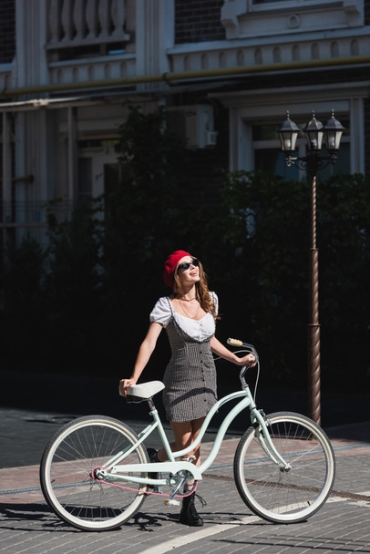 full length of happy woman sunglasses, red beret and dress standing with bicycle on street in europe  - Valokuva, kuva