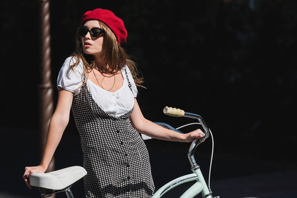 pretty young woman in sunglasses, beret and dress standing with bicycle  - Valokuva, kuva