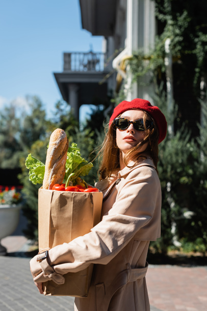 young woman in beige trench coat, sunglasses and red beret holding paper bag with groceries  - Фото, зображення