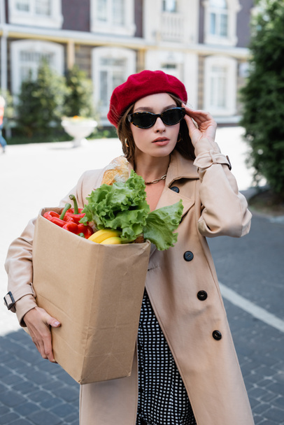 young woman in beige trench coat and beret adjusting sunglasses while holding paper bag with groceries outside - Foto, immagini