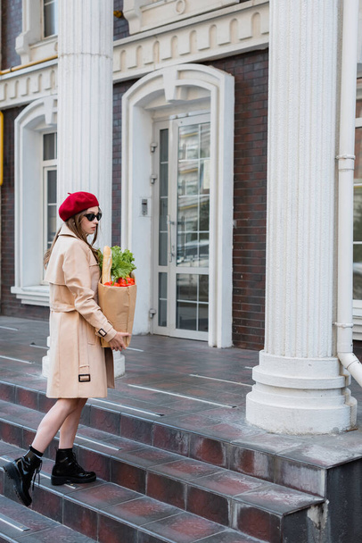in voller Länge hübsche junge Frau in Trenchcoat, Sonnenbrille und roter Baskenmütze hält Papiertüte mit Lebensmitteln in der Nähe des Gebäudes  - Foto, Bild