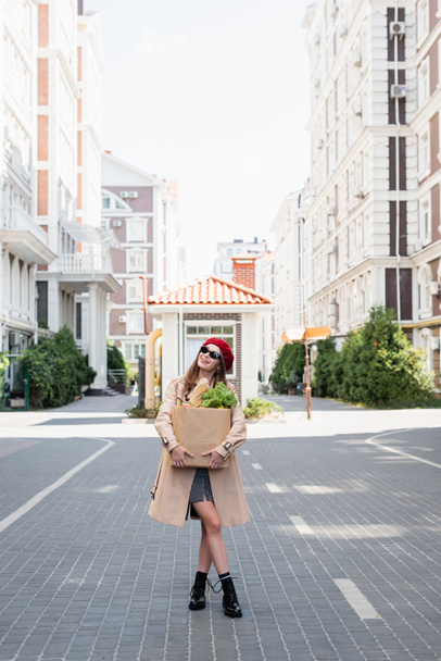 pleine longueur de femme gaie en trench coat, lunettes de soleil et béret rouge tenant sac en papier avec épicerie sur la rue urbaine  - Photo, image