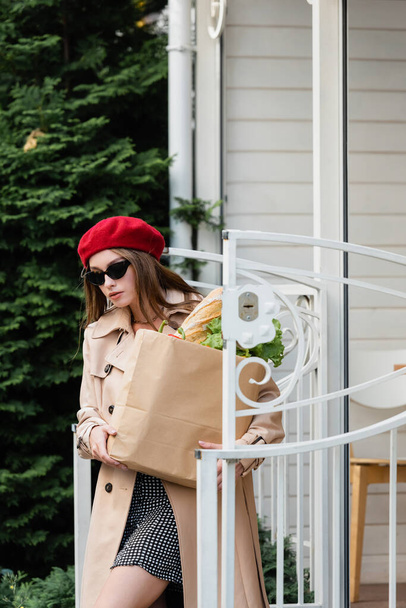 jeune femme en trench coat beige, lunettes de soleil et béret tenant sac en papier avec épicerie près du bâtiment à l'extérieur  - Photo, image