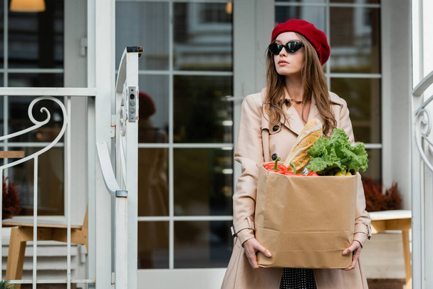 young woman in beige trench coat, red beret and sunglasses holding paper bag with fresh groceries near house  - Photo, Image