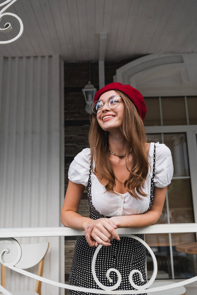 happy young woman in red beret and eyeglasses on balcony of house  - Foto, Imagem