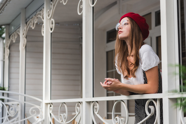 young woman in red beret and eyeglasses standing on balcony and looking away - Foto, afbeelding