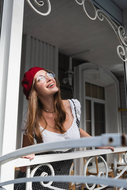 cheerful young woman in red beret and eyeglasses standing on balcony near house  - Foto, Imagem