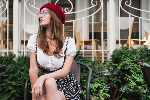 young woman in red beret, dress and eyeglasses sitting on chair outside - Foto, immagini