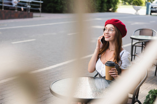 Lächelnde Frau mit roter Baskenmütze und Brille hält Pappbecher in der Hand und spricht auf der Terrasse des Cafés mit dem Smartphone - Foto, Bild