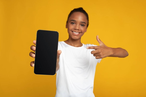 Cheerful teen afro american female pupil in white t-shirt points finger at smartphone with blank screen - Fotoğraf, Görsel