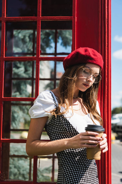 young woman in beret and eyeglasses holding paper cup near red phone booth - Фото, изображение