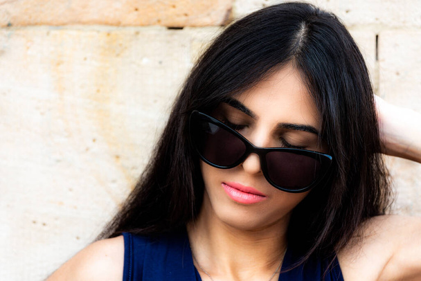 Junge dunkelhaarige Frau mit gesenkter Sonnenbrille auf der Nase posiert mit geschlossenen Augen und einem gelassenen Gesichtsausdruck, während sie einen Moment allein genießt - Foto, Bild