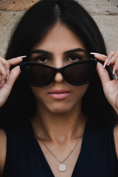 Serious young woman with a thoughtful intense expression lowering her sunglasses to stare at the camera in a close up cropped head and shoulders portrait - Photo, Image