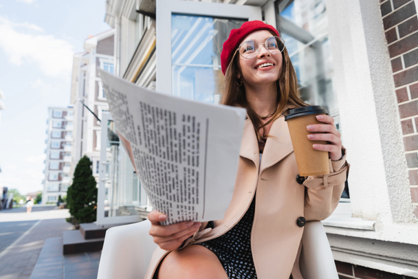 pleased young woman in red beret and beige trench coat holding newspaper and paper cup - 写真・画像