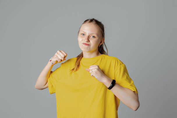 Teini-ikäinen tyttö, jolla on negatiivinen ilme, nostaa nyrkkejä raivokkaasti katsomalla kameraan, eristettynä vaaleanharmaalla taustalla, yllään rento tyhjä keltainen t-paita. Raivo ja aggressiivinen käsite - Valokuva, kuva