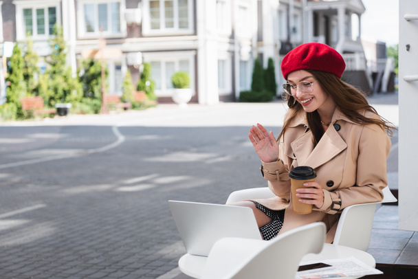 happy young woman in red beret and beige trench coat waving hand during video call on terrace of cafe  - Photo, Image