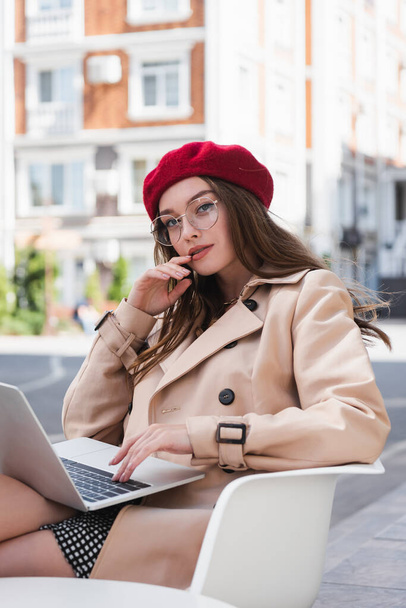 pretty freelancer in red beret and beige trench coat using laptop on terrace of cafe  - Photo, Image