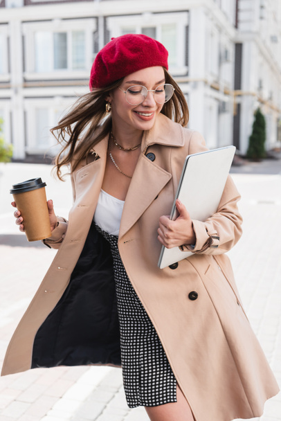 cheerful freelancer in red beret and beige trench coat holding laptop and paper cup of street  - Valokuva, kuva