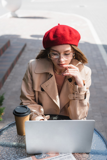 pretty freelancer in eyeglasses, red beret and beige trench coat using laptop near paper cup on terrace of cafe  - Photo, image
