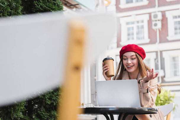 Glückliche Freiberuflerin in roter Baskenmütze hält Pappbecher in der Hand und gestikuliert beim Videoanruf im Café - Foto, Bild