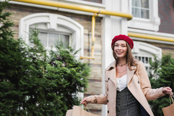 young happy woman in red beret and beige trench coat holding shopping bags outside - Fotó, kép
