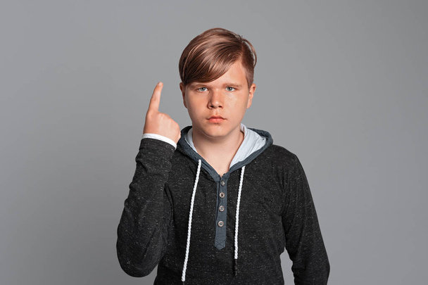 Seriöser Teenie-Junge mit erhobenem Zeigefinger, blickt in die Kamera, steht vor grauem Hintergrund, lässig gekleidet. Ausdrucksstarke Mimik - Foto, Bild